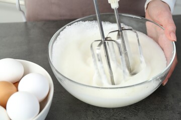 Woman beating egg whites with mixer at grey table in kitchen, closeup