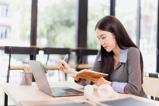Asian woman studying at a modern home workspace, using a laptop and holding a pencil over an open notebook, focused on e-learning, planning and note-taking - Powered by Adobe