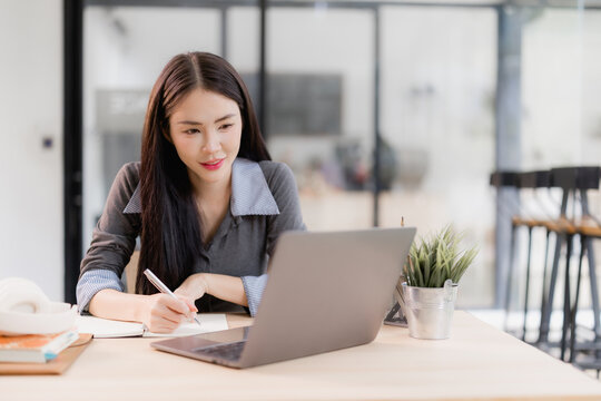 Asian student or businesswoman working on a laptop and writing notes in a notebook on a wooden desk, emphasizing remote work, distance education, or hybrid learning concepts in a modern office setup