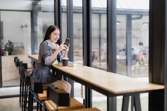 Asian woman sitting at a coffee shop bar counter, focused on her smartphone with headphones around her neck, enjoying a moment of connection or study while a coffee cup and book rest nearby
