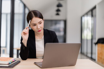 Asian businesswoman focusing on a laptop, holding a pen to her chin, portraying thoughtfulness, problem-solving, and concentration in a professional office environment