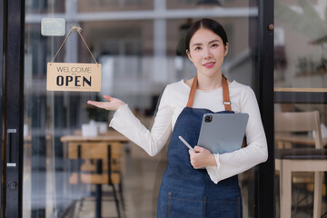 Young Asian woman cafe owner wearing an apron, holding a tablet, standing by a glass door with an OPEN sign, welcoming new customers to her small business