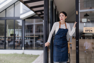 Fototapeta premium Young Asian woman small business owner wearing an apron, opening the glass door of her coffee shop with an open sign, ready to welcome customers for the day