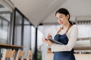 Young Asian woman barista wearing an apron standing in a cafe, confidently using a digital tablet with a stylus, taking customer orders, and managing daily operations