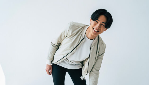 純白のスタジオ背景に立つ20代の日本人男性のポートレート。白いTシャツにライトベージュのボンバージャケットを重ね、黒のスリムパンツで引き締めたシンプルで都会的なスタイル。短い黒髪と明るくエネルギッシュな笑顔、少し前傾した躍動感ある姿勢が、爽やかで清潔感のあるムードを演出。