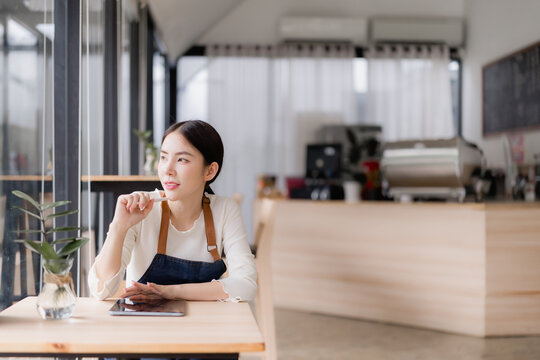Young Asian cafe owner in apron sits at a table with a tablet, gazing thoughtfully out the window during a quiet break, planning and dreaming about her business future