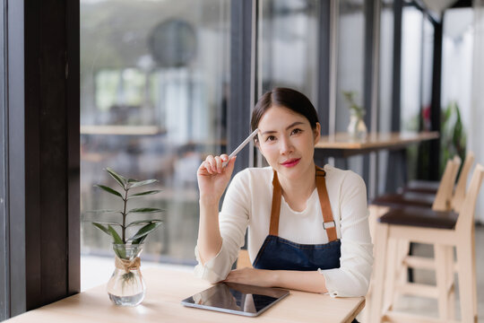 Asian coffee shop owner in an apron holds a stylus at a wooden table with a tablet, thoughtful and confident, embodying small business entrepreneurship and digital innovation