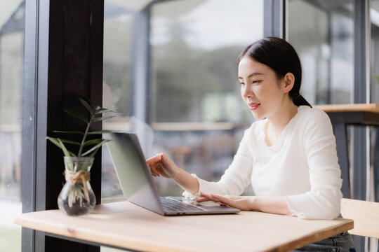 Young Asian woman smiling while remote working on a laptop at a wooden table in a modern cafe, symbolizing flexibility, freelance work, and digital nomad lifestyle