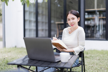 Asian woman smiling while remote working outdoors, sitting by a laptop and writing in a notebook, demonstrating flexibility and modern work-life balance in a home garden setting