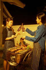 Two Asian women in striped aprons toast with glasses or beer bottles during a backyard night BBQ. Skewered marinated meats grill densely, creating a warm,festive atmosphere of friendship celebration