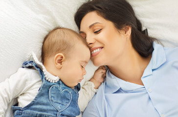 Happy smiling young mother lying in bed next to peacefully sleeping sweet baby during daytime sleep. Loving mom watching her cute sleeping newborn child and enjoying warm tender moment of closeness.