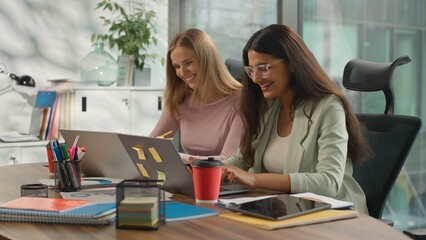 Two multiethnic women discuss graphic statistics corporate growth in office diverse businesswomen informal business girls Caucasian Indian ladies working online laptops together talking happy smiling