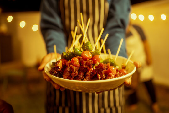 An Asian woman enjoys a cozy backyard BBQ, preparing or serving skewered meats and vegetables. Warm golden lights create a festive, relaxed atmosphere, highlighting friendship, celebration, outdoor