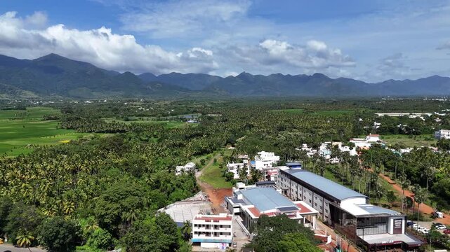 A sweeping aerial view captures a South Indian transitional landscape. Modern commercial development juxtaposes with immense coconut palm groves and traditional settlements. A majestic mountain ranges