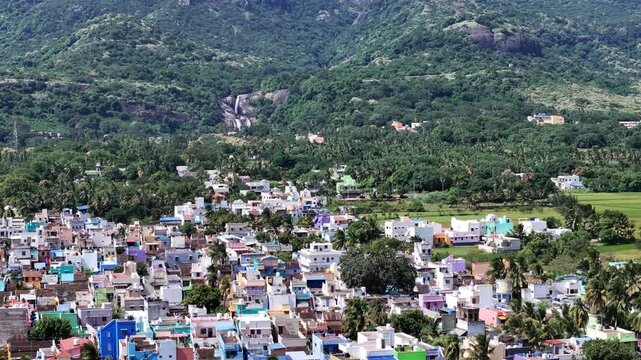 A majestic famous Courtallam (Kutralam) Main Falls in Tamil Nadu, India. cascades down a rocky, forested mountainside. A settlement is nestled below in the lush valley.