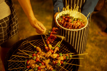 An Asian woman enjoys a cozy backyard BBQ, preparing or serving skewered meats and vegetables. Warm golden lights create a festive, relaxed atmosphere, highlighting friendship, celebration, outdoor