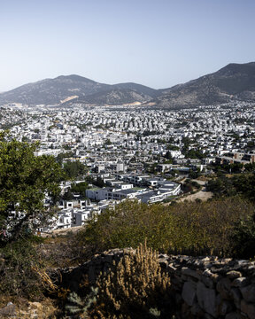 View of white buildings cascading down the hillside towards the shimmering blue sea, nestled between rugged mountains under a clear sky, Bodrum, Mugla, Turkey.
