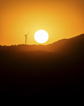 View of a lone wind turbine stands silhouetted against the fiery sunset over rolling hills, casting long shadows in Bodrum, Mugla, Turkey.