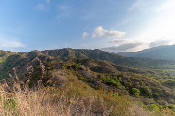 A wide landscape view of a vast mountain valley. The valley is filled with green bushes. The sun is bright, creating a hazy atmosphere.
