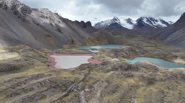 Aerial moves toward three lakes - Oqe Qocha, Puca Cocha, and Azul Cocha - connected by a dirt path, approaching the snow-capped peak of Nevado Ausangate