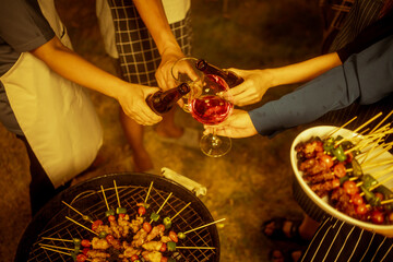 An Asian woman and friends toast with wine during a cozy backyard BBQ. Grilling skewers over charcoal under string lights, they celebrate friendship, joy,  togetherness in a festive outdoor evening