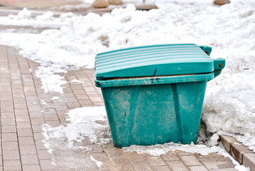Fototapeta premium Green grit box filled with sand and salt mixture placed on snowy city sidewalk for road and pedestrian safety during winter season, urban maintenance equipment. Grit container on city sidewalk