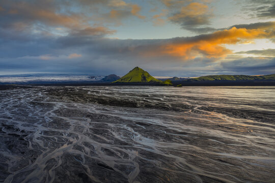 Aerial view of braided river patterns cut through the volcanic landscape under an orange-tinged sky near a distant mountain, Highlands Of Iceland, Sveitarf&eacute;lagio Hornafjorour, Iceland.