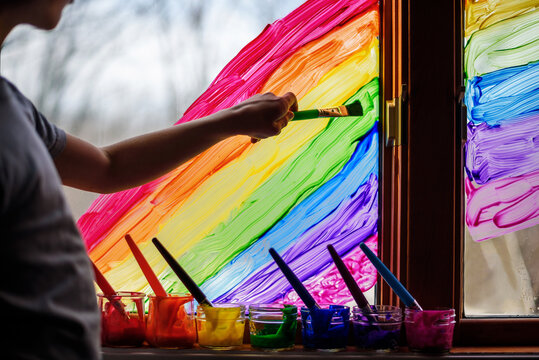 Close-up side view of a Boy painting a rainbow on a window with colourful acrylic paint