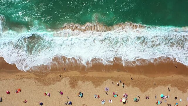 Aerial drone view of tourists relaxing under parasols on sandy seashore, with waves splashing on the coast of a popular beach destination on a sunny day