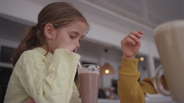 Girl in yellow sweater drinking chocolate beverage from tall glass at indoor cafe table. Another child sitting nearby. Warm and cozy setting with soft lighting and casual dining atmosphere