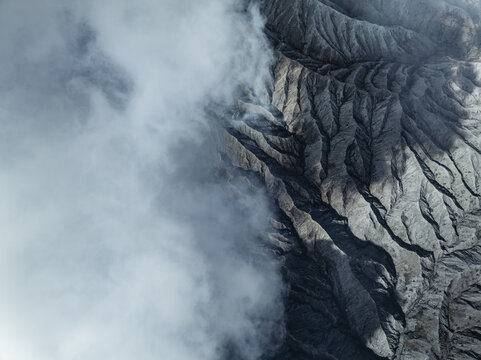 Aerial view of the rugged, textured landscape of Mount Bromo juxtaposed against ethereal clouds, creating a dramatic contrast of light and shadow, Mount Bromo, East Java, Indonesia.