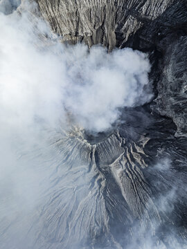 Aerial view of the majestic Mount Bromo crater exhaling ethereal white smoke against the dark, rugged terrain, East Java, Indonesia.