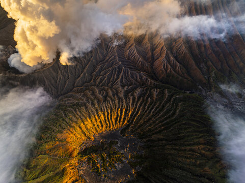 Aerial view of Mount Bromo's caldera shrouded in ethereal clouds, its volcanic slopes etched with deep ravines and bathed in golden sunlight, Mount Bromo, East Java, Indonesia.