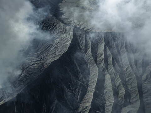 Aerial view of the rugged, grey volcanic landscape, shrouded in ethereal mist, creating a dramatic and otherworldly scene, Mount Bromo, East Java, Indonesia.