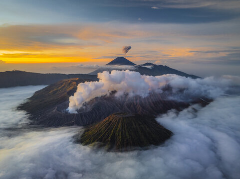 Aerial view of Mount Bromo piercing through a sea of clouds at dawn, with volcanic smoke painting the sky, Mount Bromo, East Java, Indonesia.