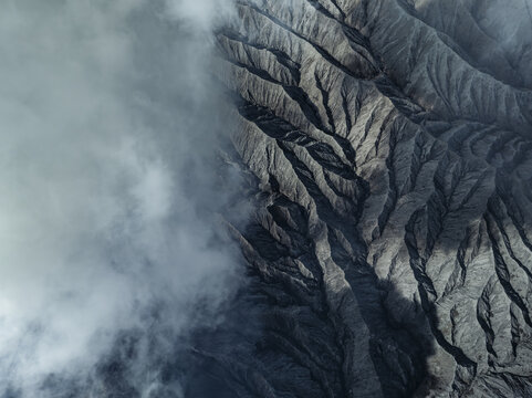 Aerial view of a rugged volcanic landscape shrouded in ethereal mist, revealing stark contrasts between light and shadow on the textured terrain, Mount Bromo, East Java, Indonesia.