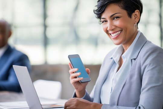 Confident businesswoman smiling at camera while holding smartphone and working on laptop in office. Professional woman using technology for communication, networking or productivity.