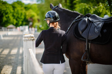 A young female jockey stands next to her black horse, wearing a special uniform and helmet, against a blue sky on a sunny summer day. View from behind. Equestrian competition and event.