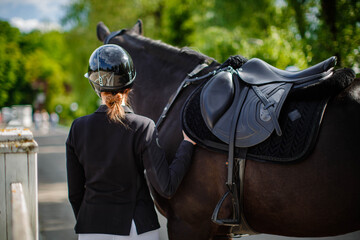 A young female jockey stands next to her black horse, wearing a special uniform and helmet, against a blue sky on a sunny summer day. View from behind. Equestrian competition and event.