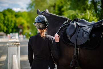 A young female jockey stands next to her black horse, wearing a special uniform and helmet, against a blue sky on a sunny summer day. View from behind. Equestrian competition and event.