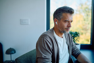 Obraz premium Middle-aged man wearing earbuds, relaxing on a sofa, and enjoying private audio content by a large window