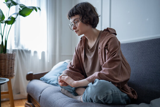 Anxious upset woman sitting on sofa at home nervously fidgeting with hands. Stressed troubled young female feeling worried, tense suffering from intrusive thoughts. Depression, burnout, inner conflict