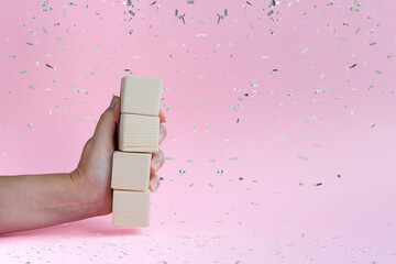 The hand holds four empty wooden dice on a pink background with silver confetti and space for text
