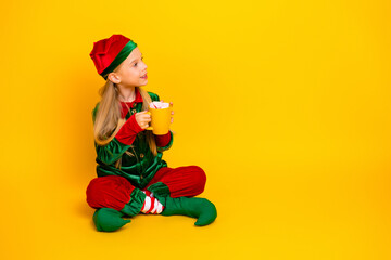 Adorable girl dressed as a festive elf in a vibrant costume sipping hot cocoa on a yellow background