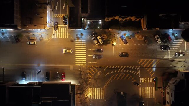 Top down aerial view of cars driving and parking on a city street at night, with bright street lights illuminating the roads and moderate traffic