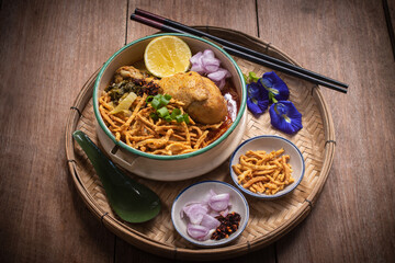 Beautiful still life of traditional Northern Thai Khao Soi chicken curry noodles served in an enamel bowl on a wooden table, with crispy noodles, shallots, lime, chili oil.