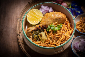 Beautiful still life of traditional Northern Thai Khao Soi chicken curry noodles served in an enamel bowl on a wooden table, with crispy noodles, shallots, lime, chili oil.