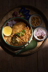 Beautiful still life of traditional Northern Thai Khao Soi chicken curry noodles served in an enamel bowl on a wooden table, with crispy noodles, shallots, lime, chili oil.