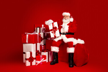 Santa Claus in a red suit with wrapped gifts sits beside a large red sack against a vivid red backdrop for a festive Christmas stock photo featuring family celebration and holiday cheer