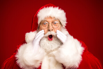 Santa Claus with surprised expression in red velvet suit and white fur trim gloved hands framing his face for a festive christmas moment